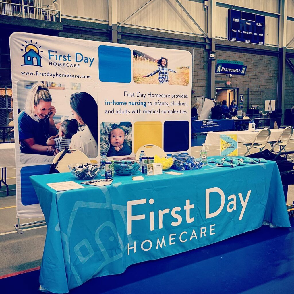 Promotional booth for First Day Homecare at an indoor event, featuring a branded table and banner highlighting in-home nursing for infants, children, and adults with medical complexities.